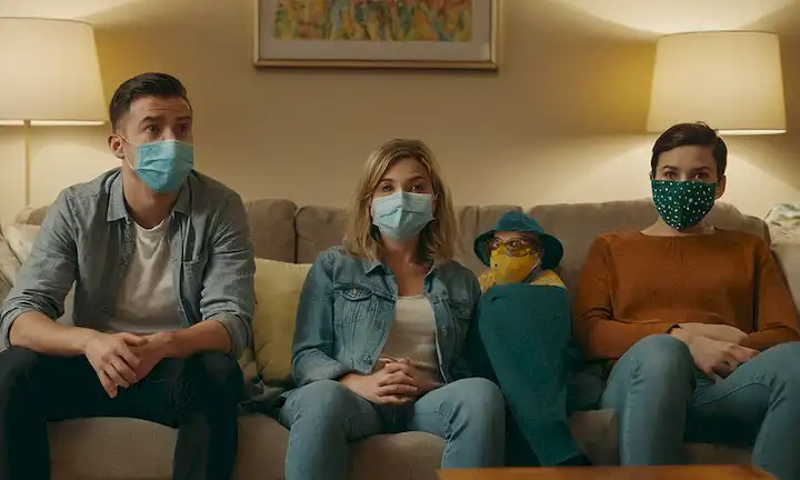 4 personas sentadas en un sofá, algunas con máscaras, otras no, con diferentes expresiones faciales, en medio de objetos y ropa dispersos, en un pequeño salón, con fondo borroso para transmitir sentido del confinamiento, y sutil insinuación de la desordenación