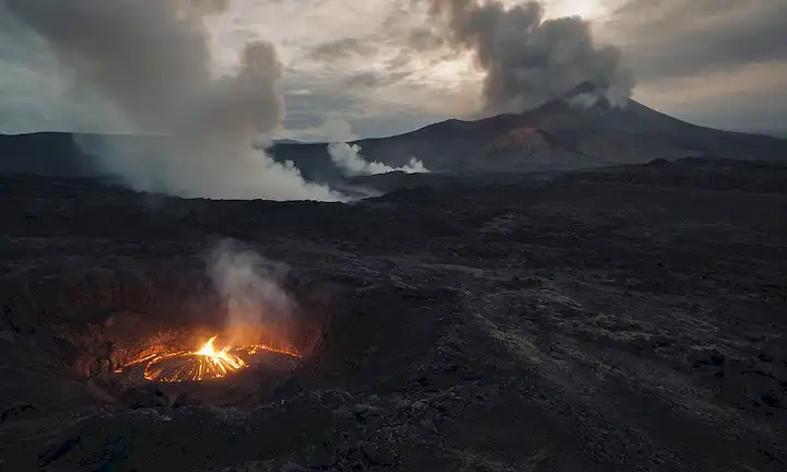 Paisaje volcánico con montañas defectuosas, cráter desconcertante en la distancia, nubes oscuras reuniéndose, vapor subiendo desde el suelo, iluminación lenceria, terreno accidentado, formaciones rocosas, wisps sutiles de gas escapando