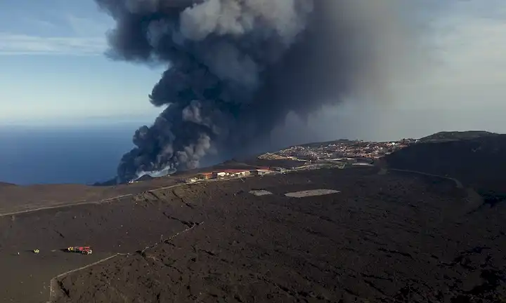 Aerial shot of La Palma islands landscape, with smoke rising from the Cumbre Vieja volcán, a crescent- shape line of destruction, fragmented rocks and ash covering the ground, dark clouds looming in the background, and emergency respondens or scientific in bormat suits in the foreground
